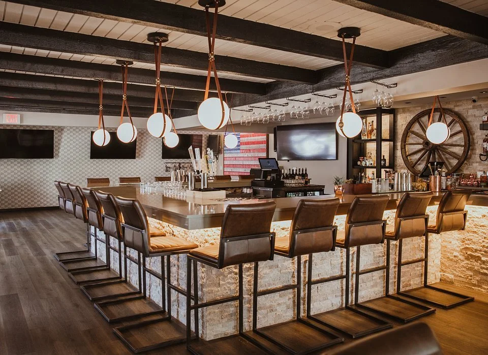 A modern bar with a stone counter, leather bar stools, hanging pendant lights, a wall-mounted TV, and shelves with bottles and glasses, in a stylish restaurant setting.