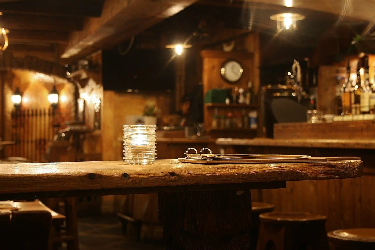 Close-up of a wooden table in a cozy bar or restaurant with a stack of coins and a pair of eyeglasses on it, warm lighting, wooden decor, and a bar counter in the background.