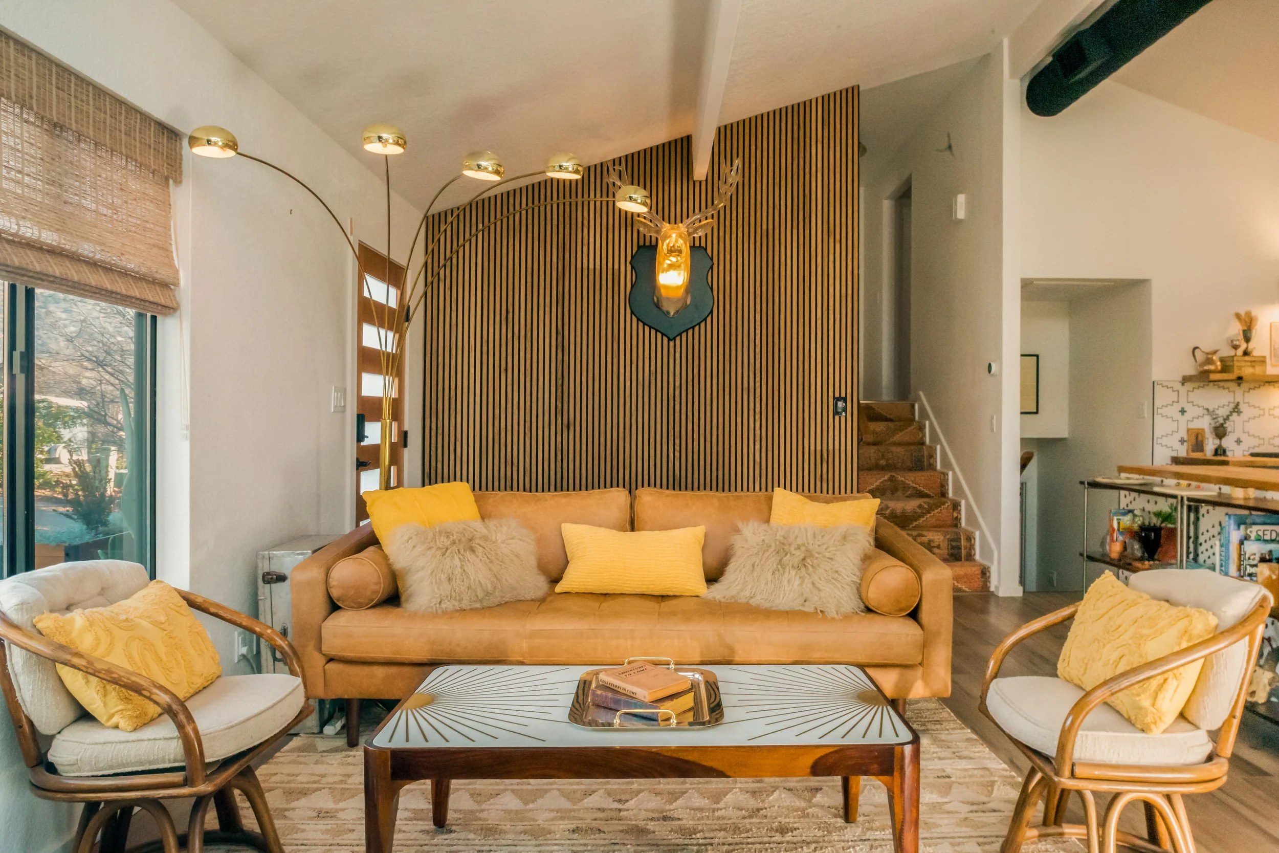 Living room with a tan sofa, yellow pillows, two armchairs with pillows, a coffee table, a wooden wall with a mounted deer head, and a window with blinds.