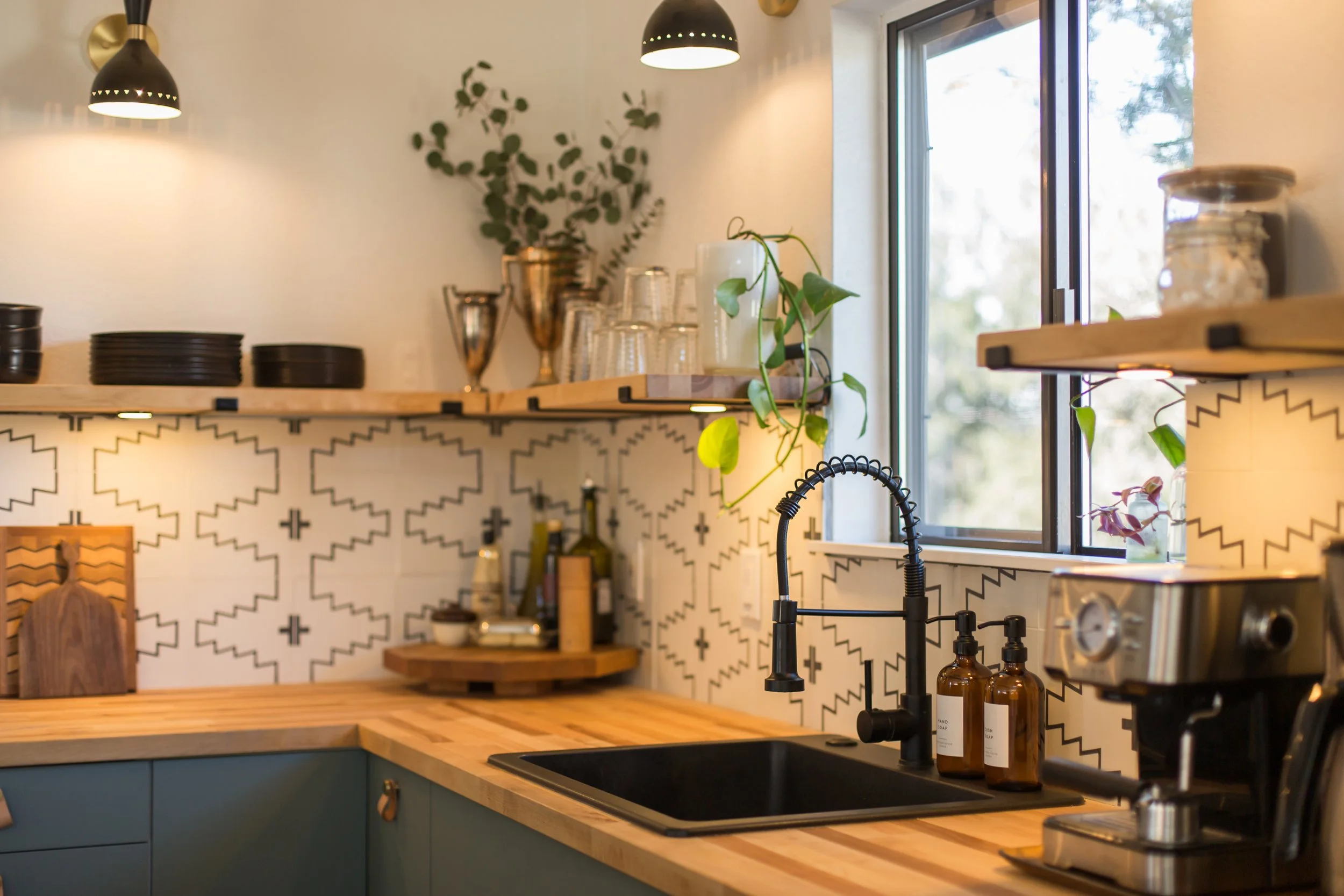 Kitchen with open window, black sink, wooden countertop, patterned tile backsplash, black faucet, bottles of soap or lotion, coffee machine, open shelves with glasses and jars, plants, and black pendant lights.
