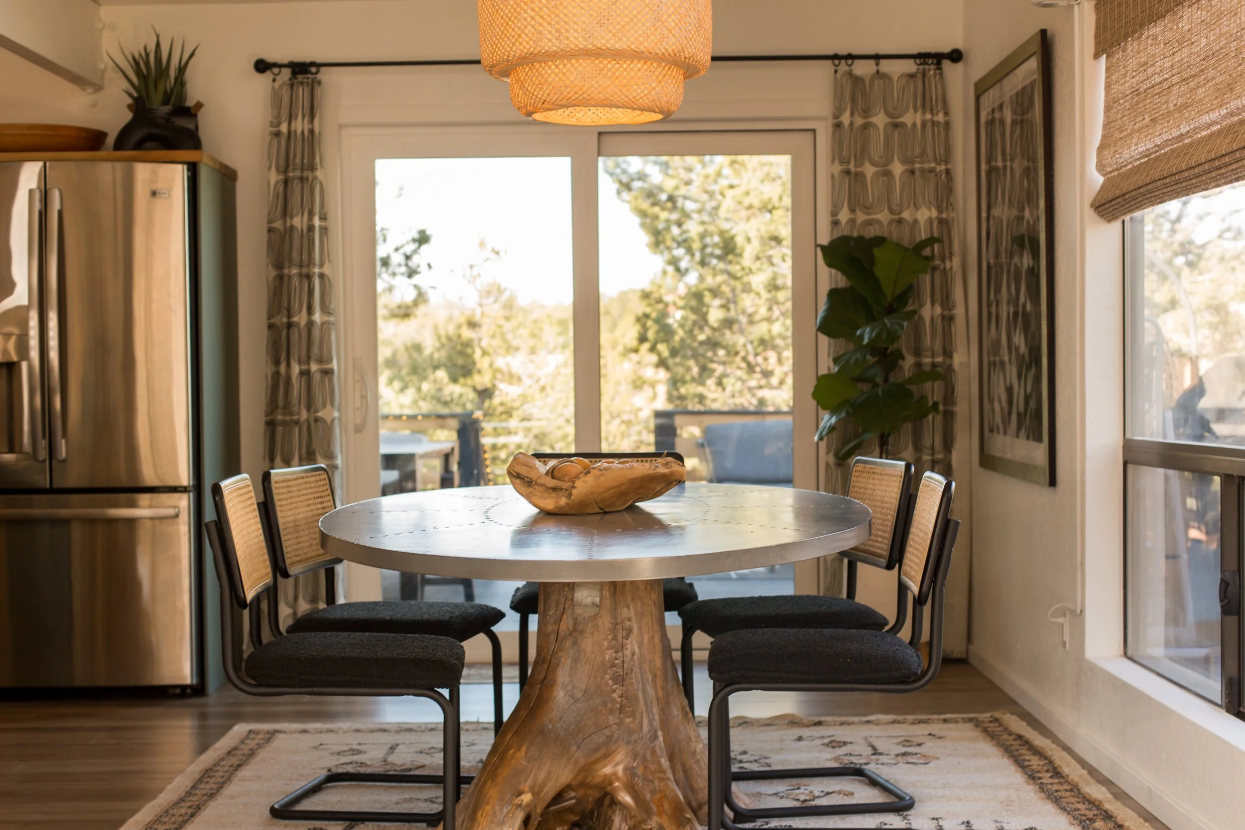 Wooden dining table with flower-shaped top, four black chairs with woven backs, a ceiling light fixture, a sliding glass door with curtains, a large plant, a framed artwork, a window with blinds, and a refrigerator in a bright dining room.