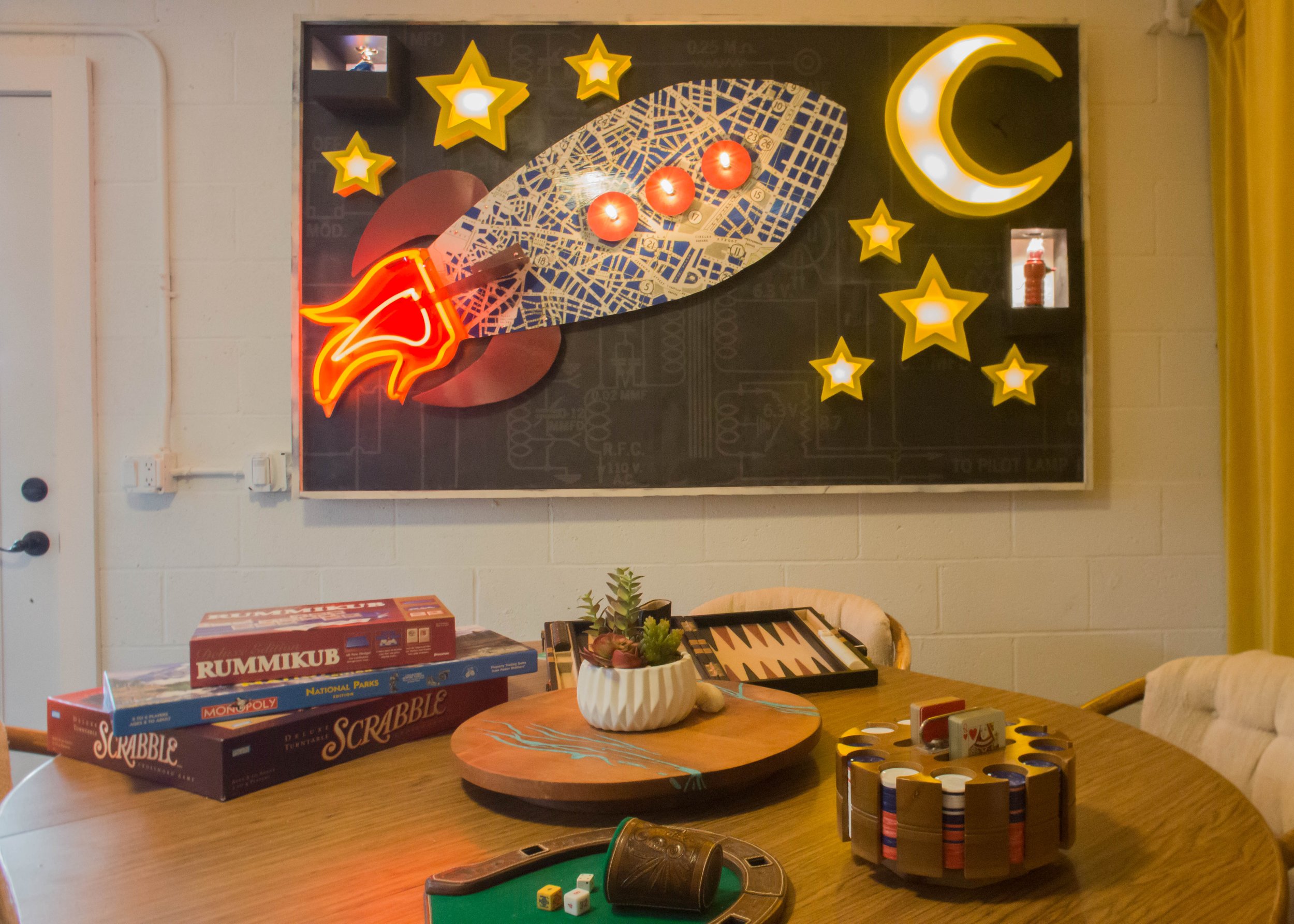 A board game table with Scrabble, Monopoly, and Rummikub boxes, a plant, and a backgammon board. A neon rocket, stars, and a crescent moon are on the wall behind.