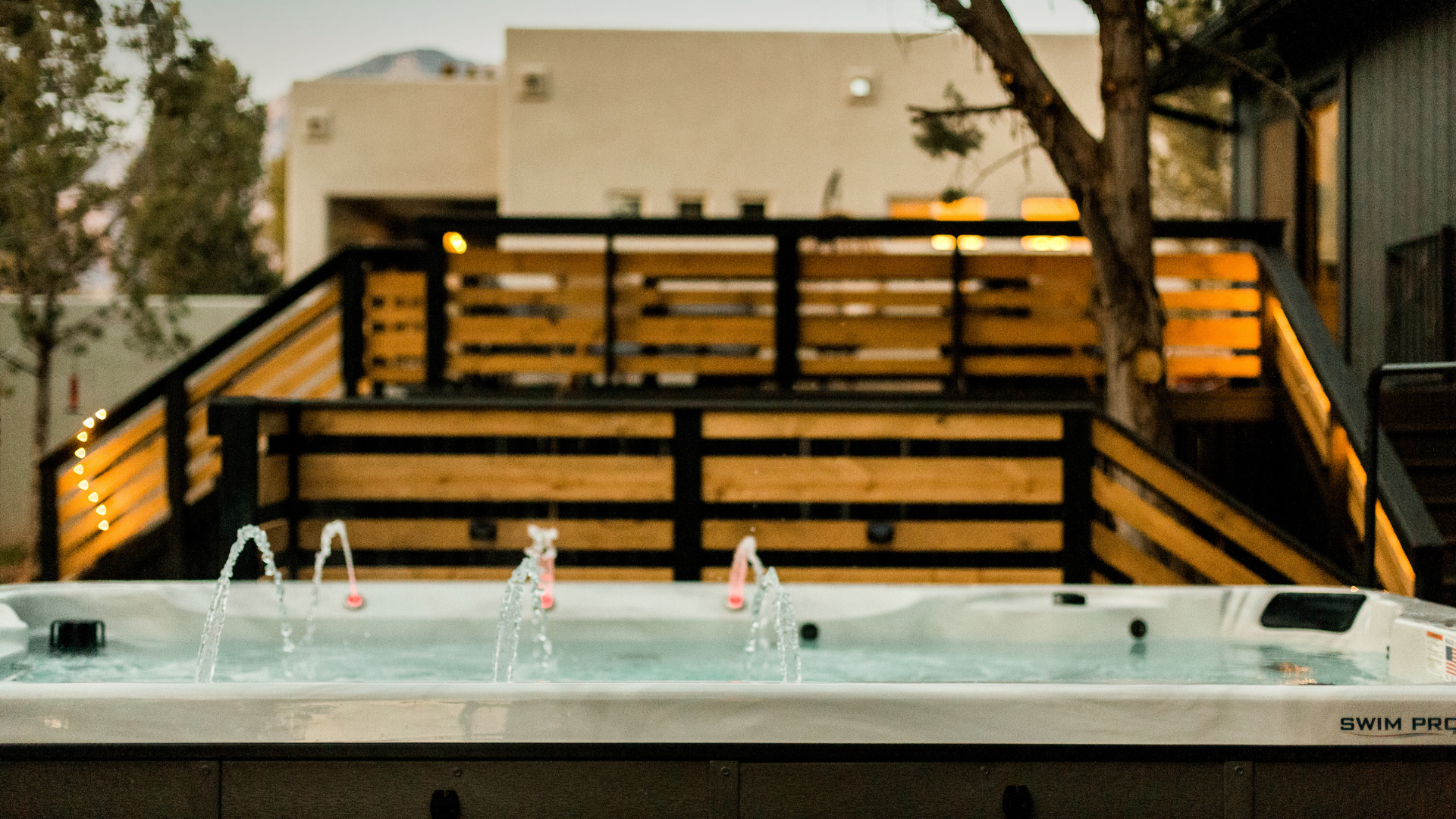 An outdoor hot tub with multiple water jets, set against a background of wooden deck stairs and nails with warm ambient lighting.