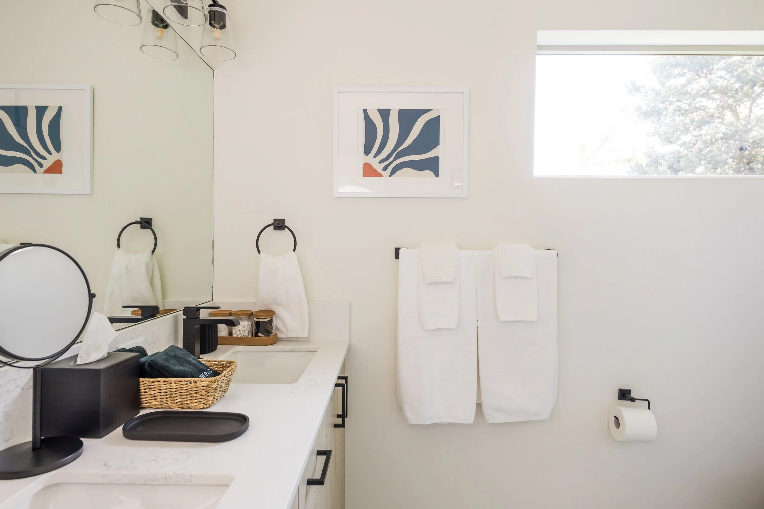 Bathroom with white walls and a large mirror, black towel rings with white towels, black and white decor, a window with sunlight, and a white countertop with toiletries.