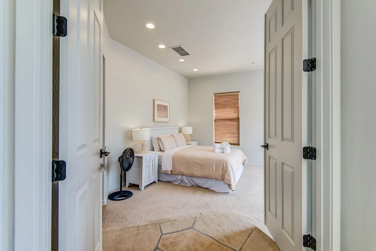 View into a bedroom through an open door, showing a bed with beige bedding, two nightstands with lamps, a window with blinds, and a small black fan
