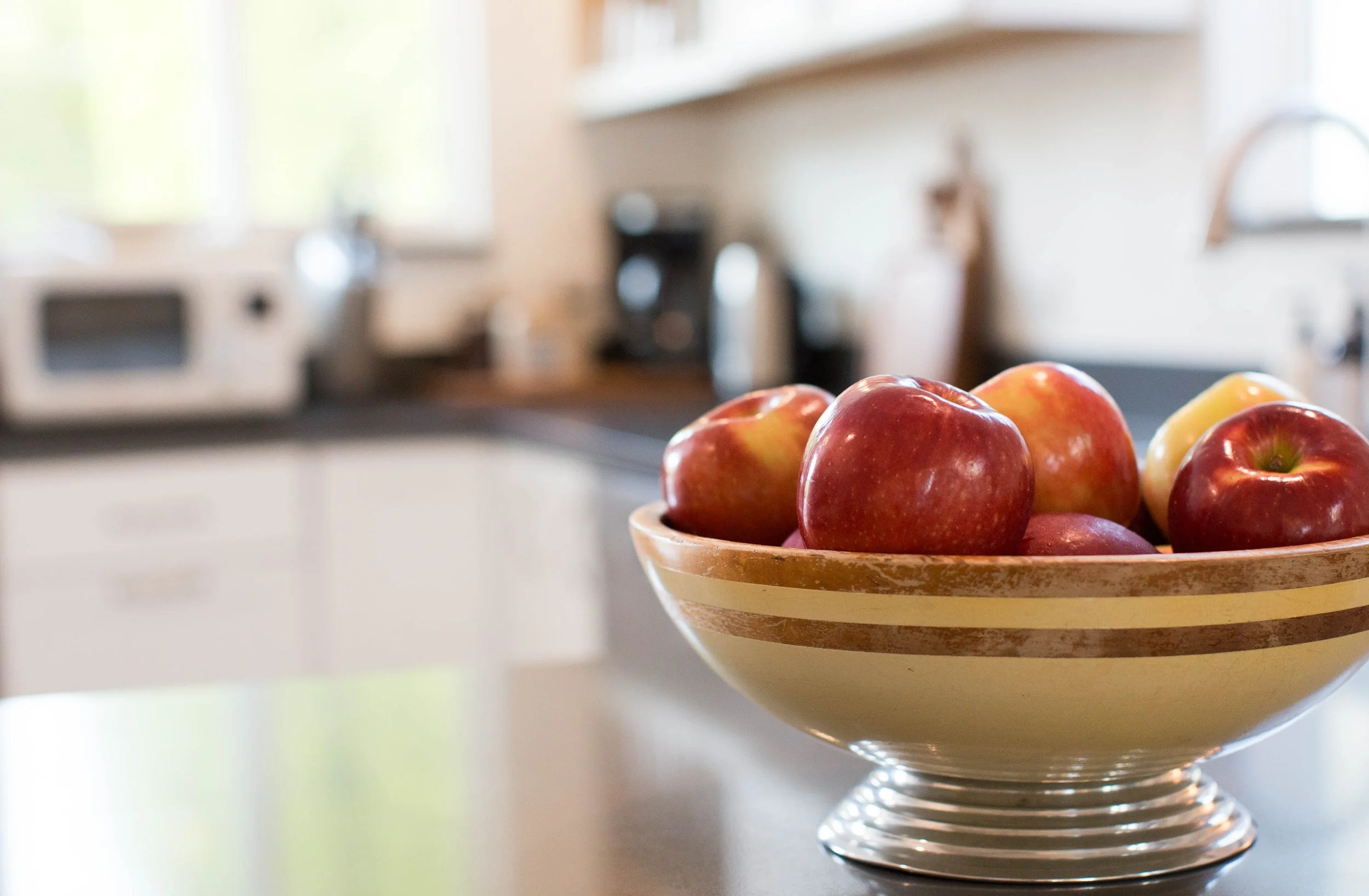 A bowl of red and yellow apples on a kitchen countertop with a blurred background of kitchen appliances and windows.