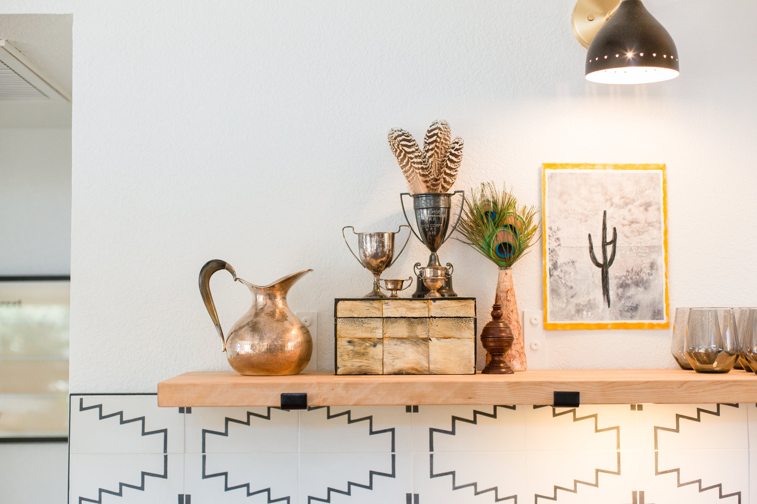 Decorative shelf with metallic pitcher, trophies, feathers, peacock feathers, framed art of cactus, and glasses on a tiled counter.
