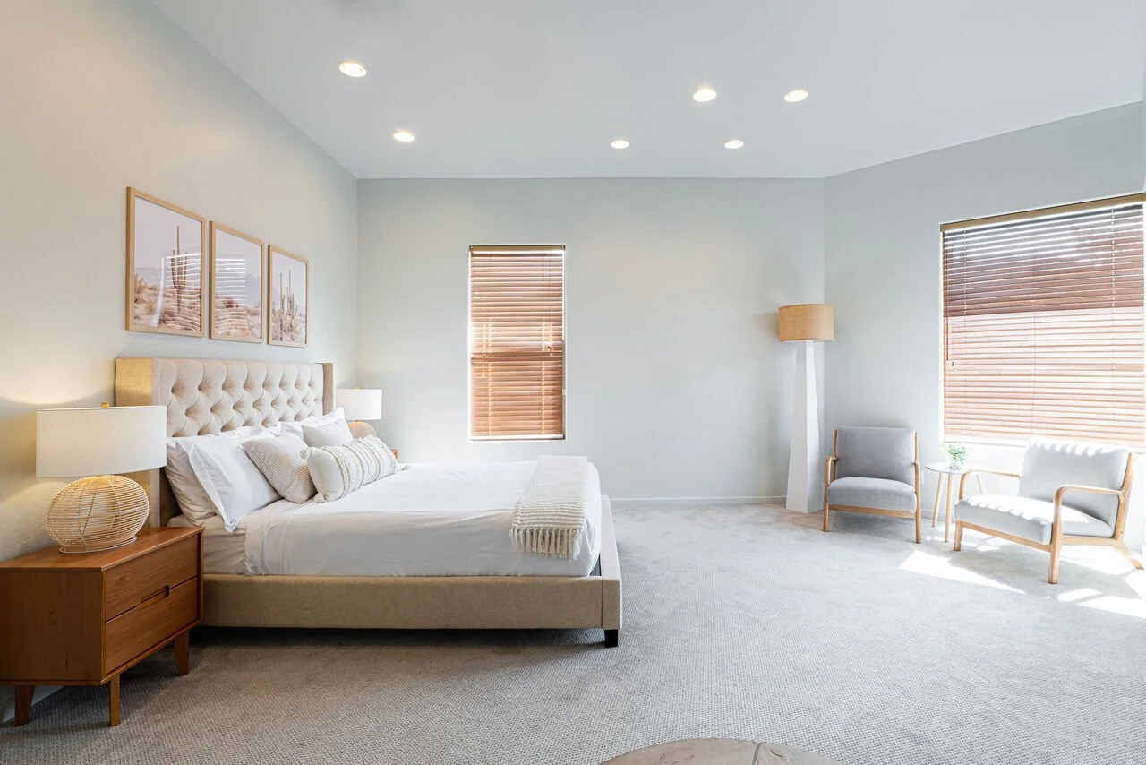 Bright, minimalistic bedroom with a large bed, wooden nightstand, and two chairs near windows with wooden blinds, and a tall floor lamp.