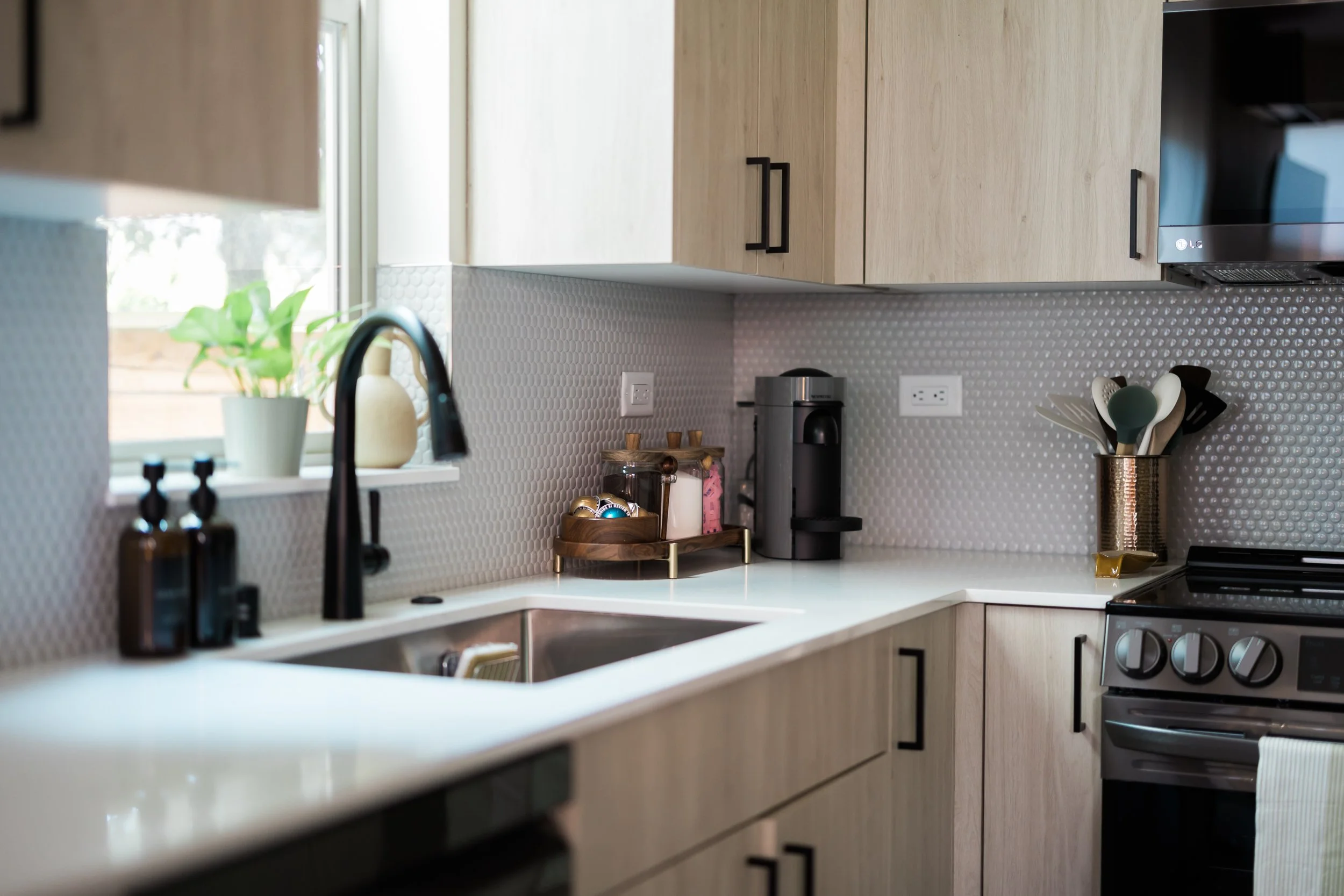 Modern kitchen with beige cabinets, a white countertop, a black kitchen sink with a black faucet, a small potted plant, soap bottles, a coffee maker, jars, a utensil holder, stovetop, and electrical outlets.