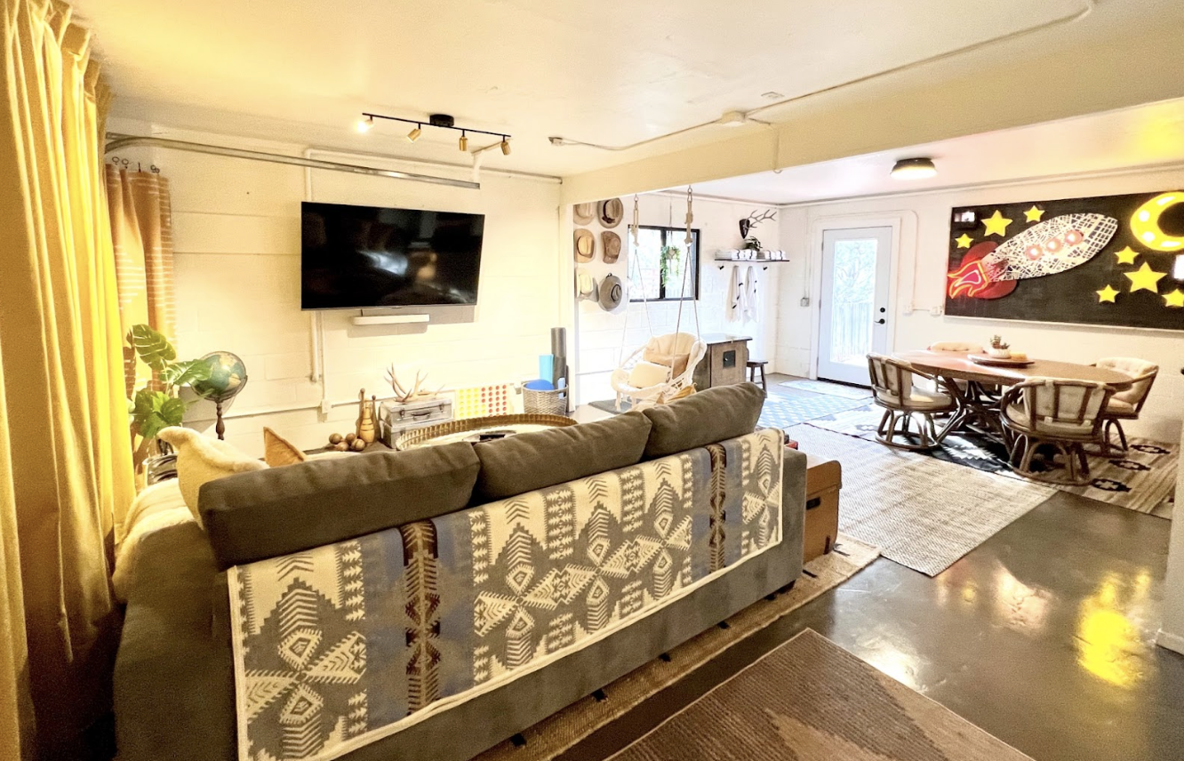 Living room with a gray sofa, a wicker chair, a wall-mounted TV, and a dining table with four chairs. Decor includes a black space-themed artwork with stars and a rocket, hats on the wall, and a swing with cushions near the window.