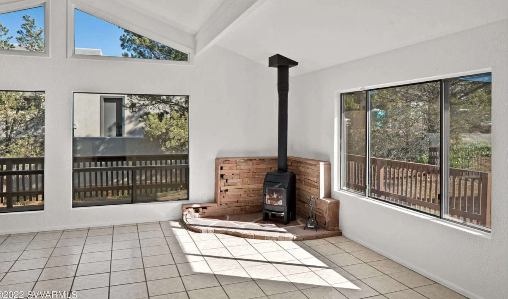 Sunlit room with large windows, white walls, tiled floor, and a wood stove with a brick surround.