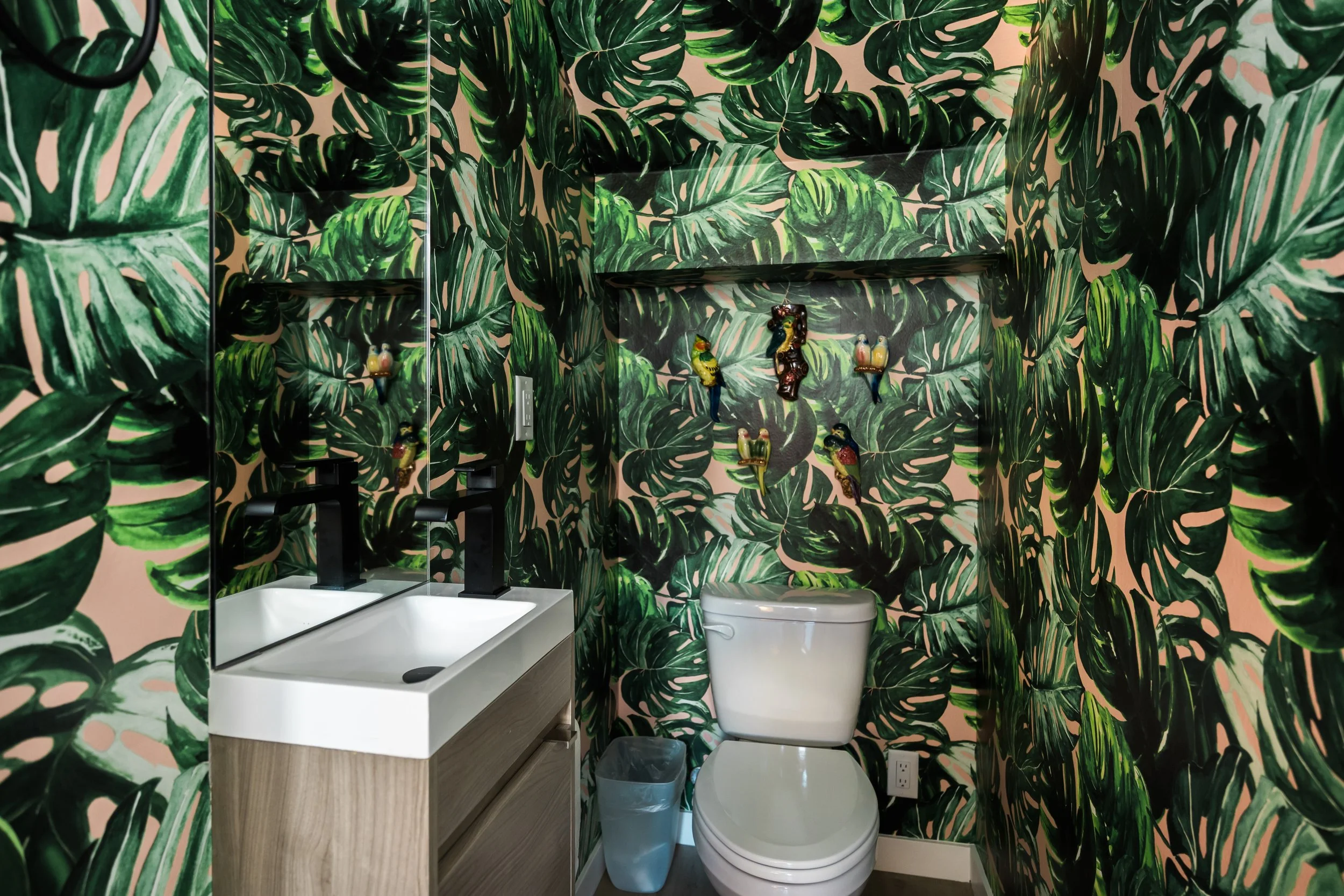 Bathroom with tropical leaf wallpaper, featuring a small vanity with a white sink, black faucet, a mirror, a toilet, and decorative bird wall hangings.
