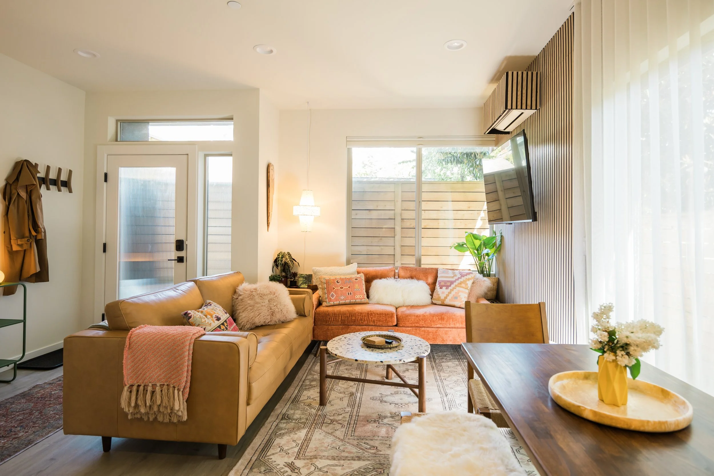 Bright living room with beige and burnt orange sofas, a round coffee table, a wooden dining table with a vase of white flowers, large windows with sheer curtains, and a sliding glass door leading outside.