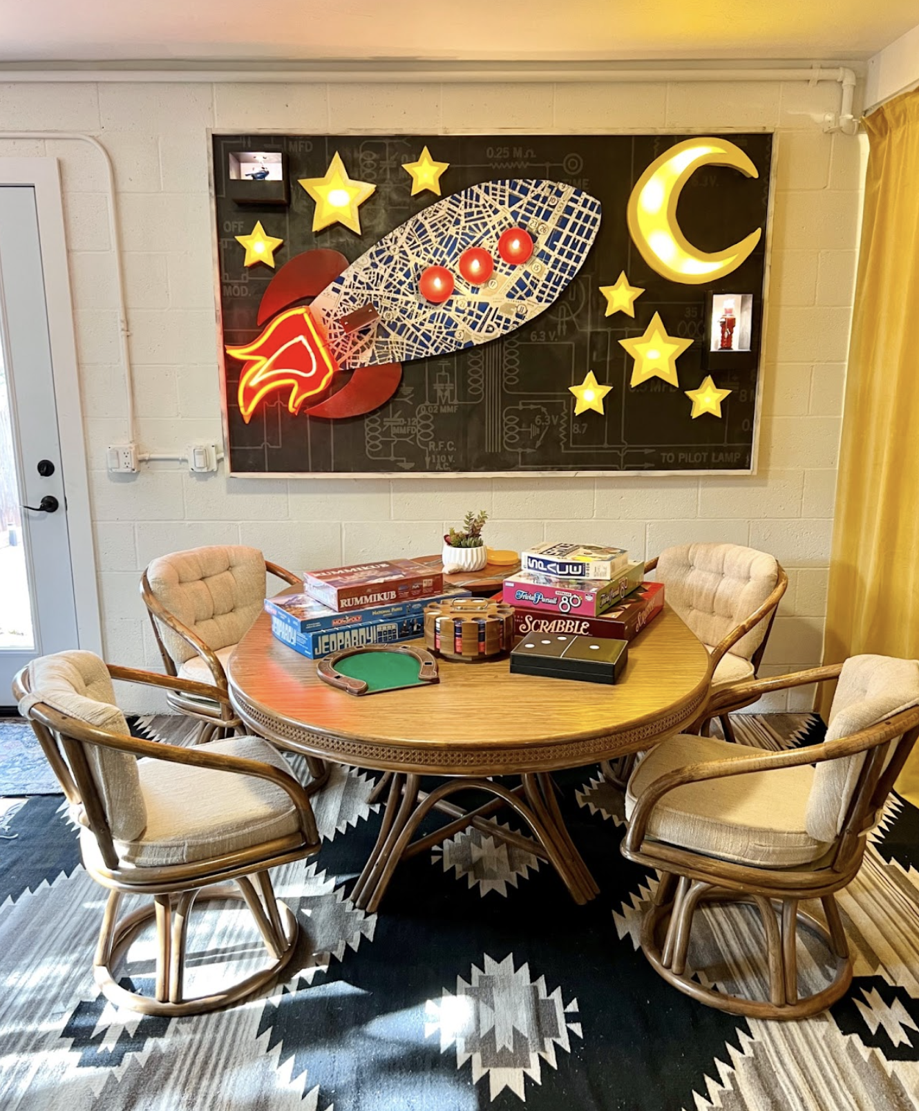 A round wooden table with six beige cushioned chairs, a board game collection including Rummikub, Scrabble, and Jumanji, and a small potted plant. Above the table is a space-themed neon wall decoration with a rocket, stars, a crescent moon, and a bla