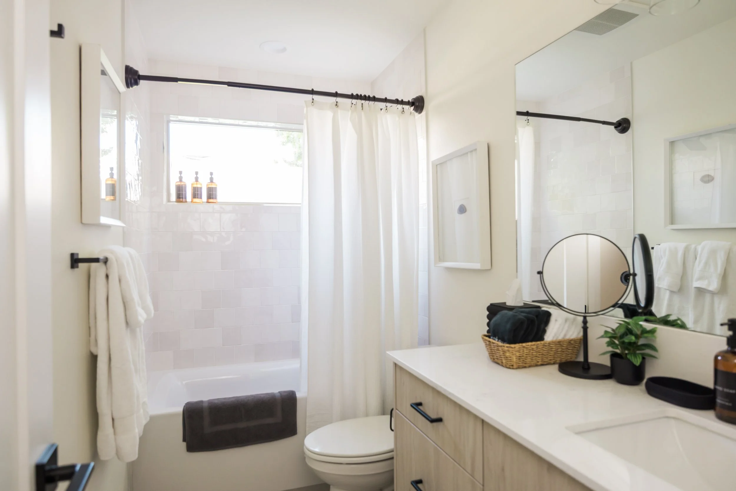 Bathroom with white counter, mirror, towels, plant, tissue box, and a shower with a window, curtain, and bottles on the ledge.
