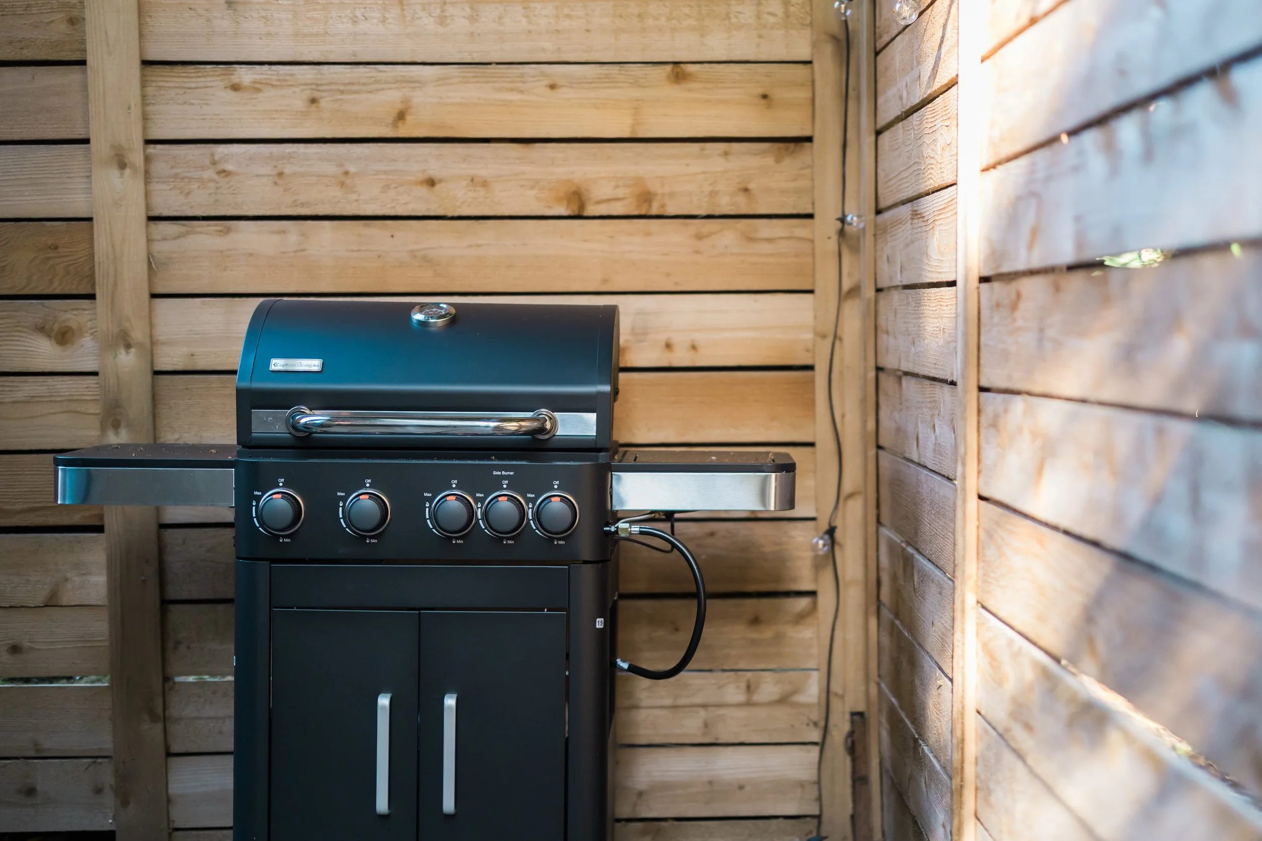 Black barbecue grill with four control knobs and a closed lid, positioned against a wooden fence wall.