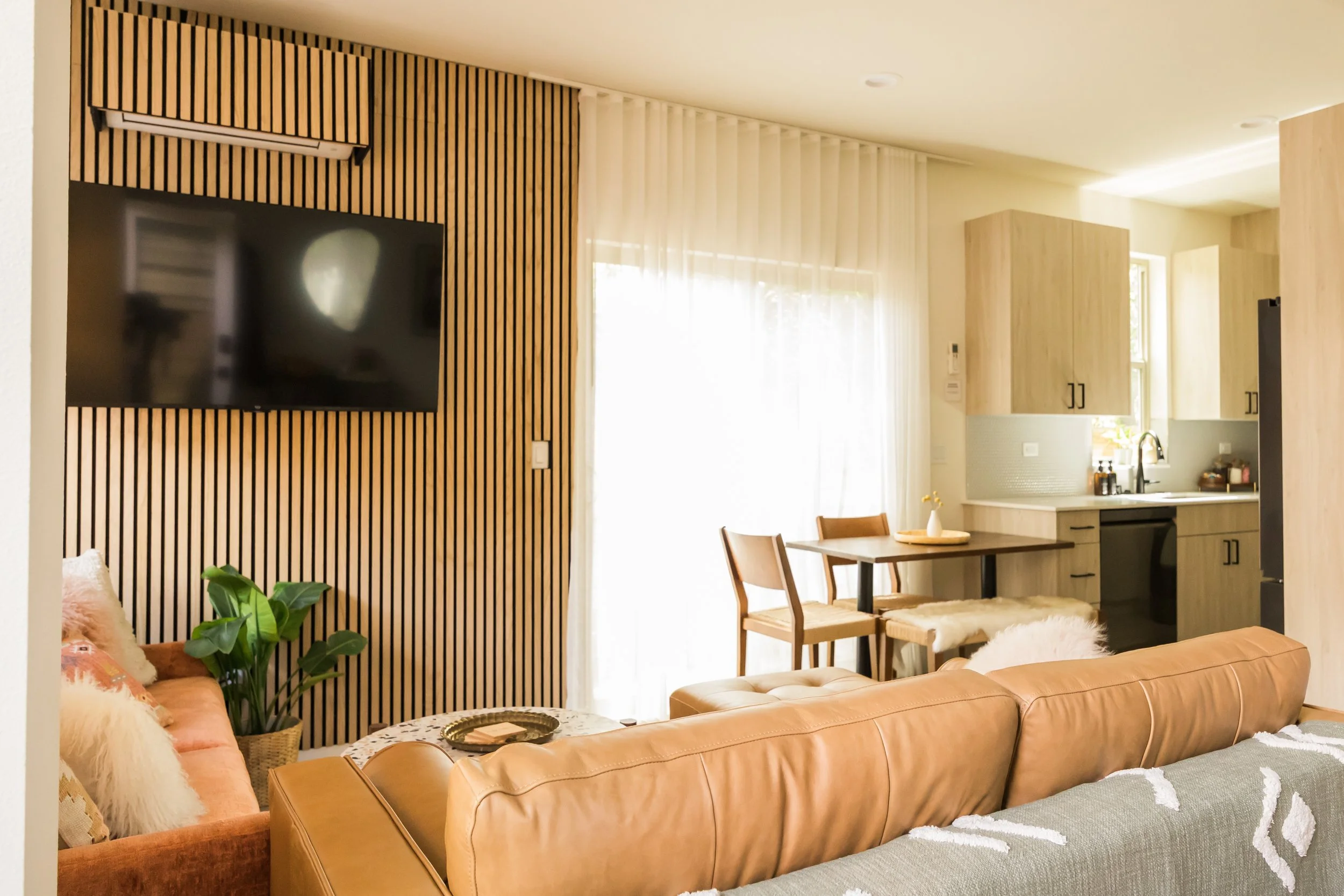 Living room with a wall-mounted TV, a tan leather sofa, a small dining table with two chairs, a kitchen area with wooden cabinets, and a window with curtains allowing sunlight.