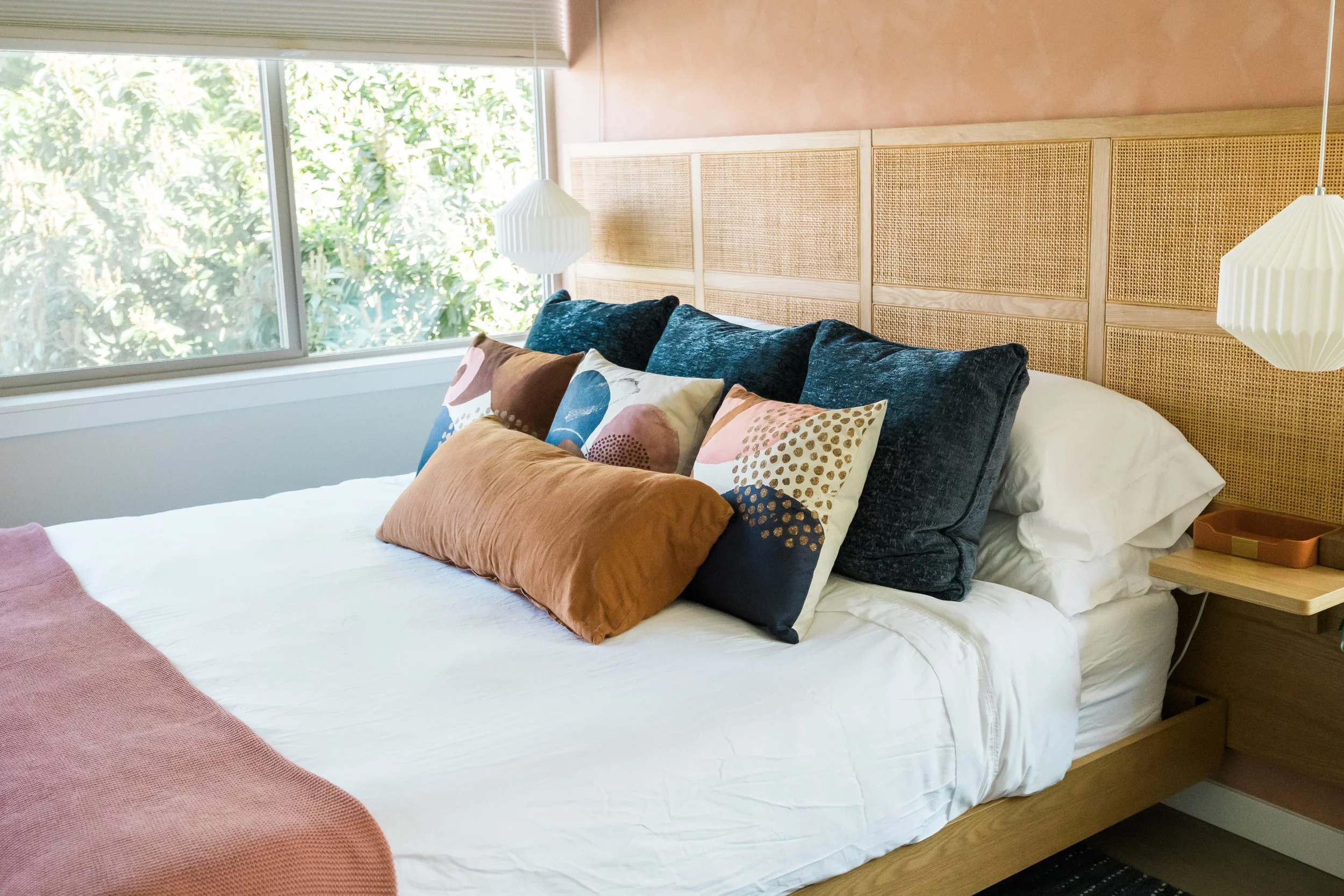 A neatly made bed with decorative pillows, a wooden headboard and a side table with a hanging lamp in a bright bedroom with a large window and greenery outside.