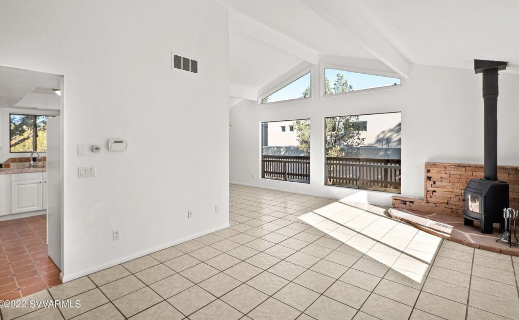 Empty living room with large windows, tiled floor, and a brick fireplace with a wood stove.