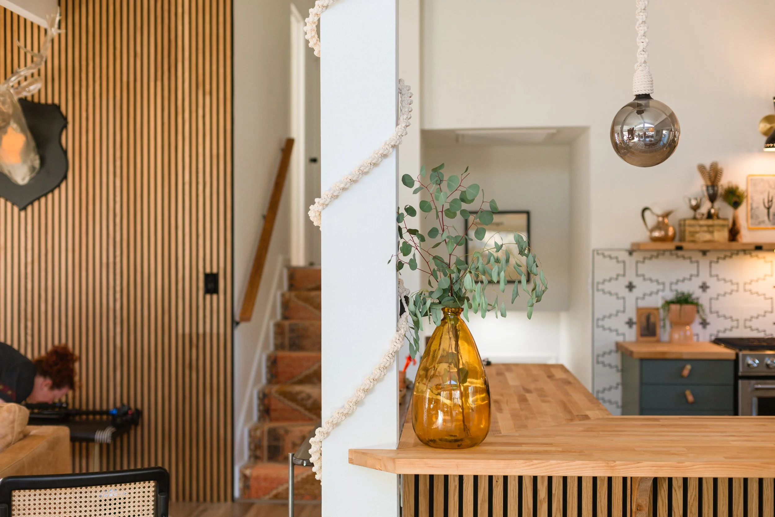 Living room and kitchen area with a wooden counter, amber vase with eucalyptus leaves, metallic hanging light, and wooden accents.