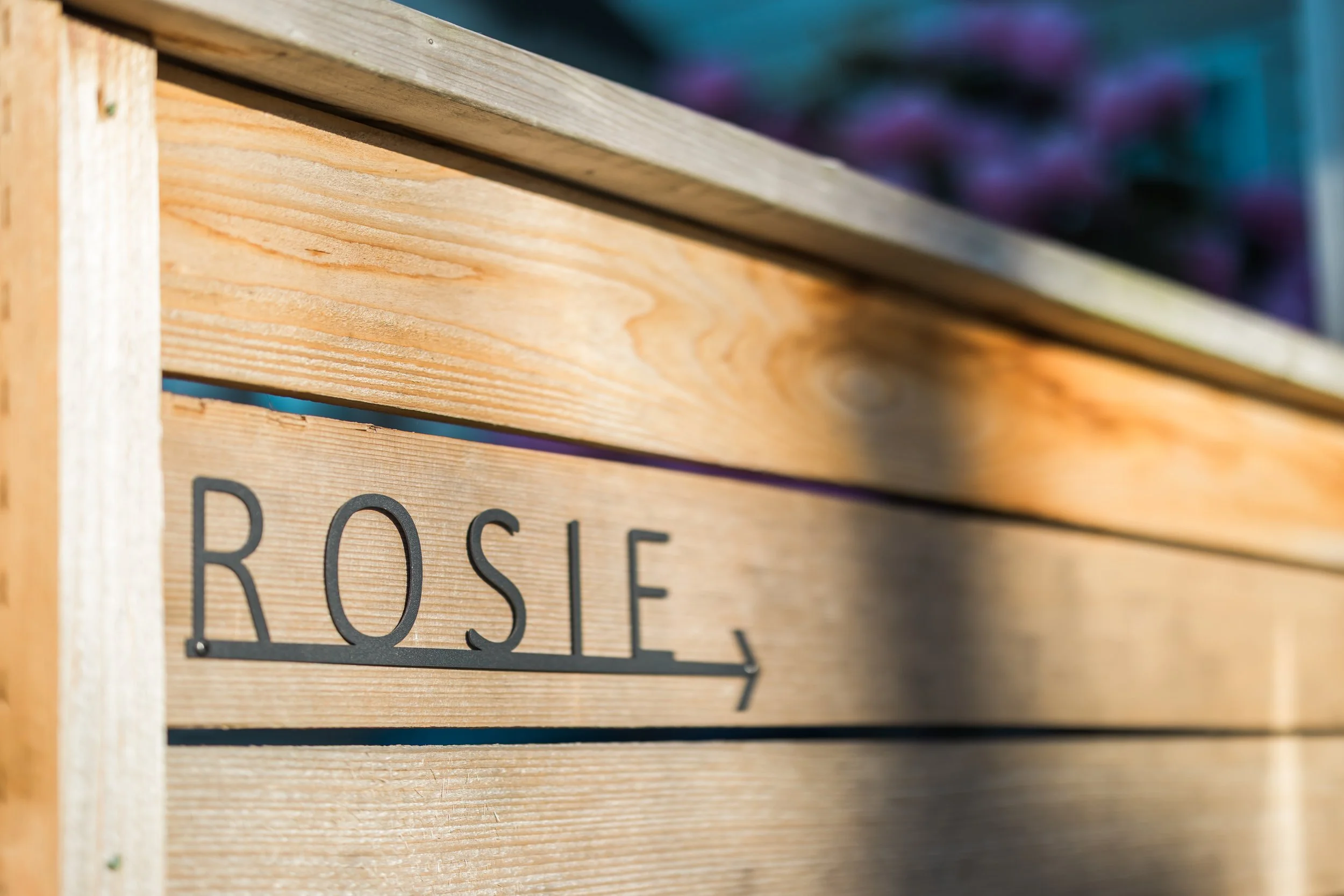 Wooden crate with the word 'Rosie' and an arrow pointing right, outdoors with blurred colorful objects in background.