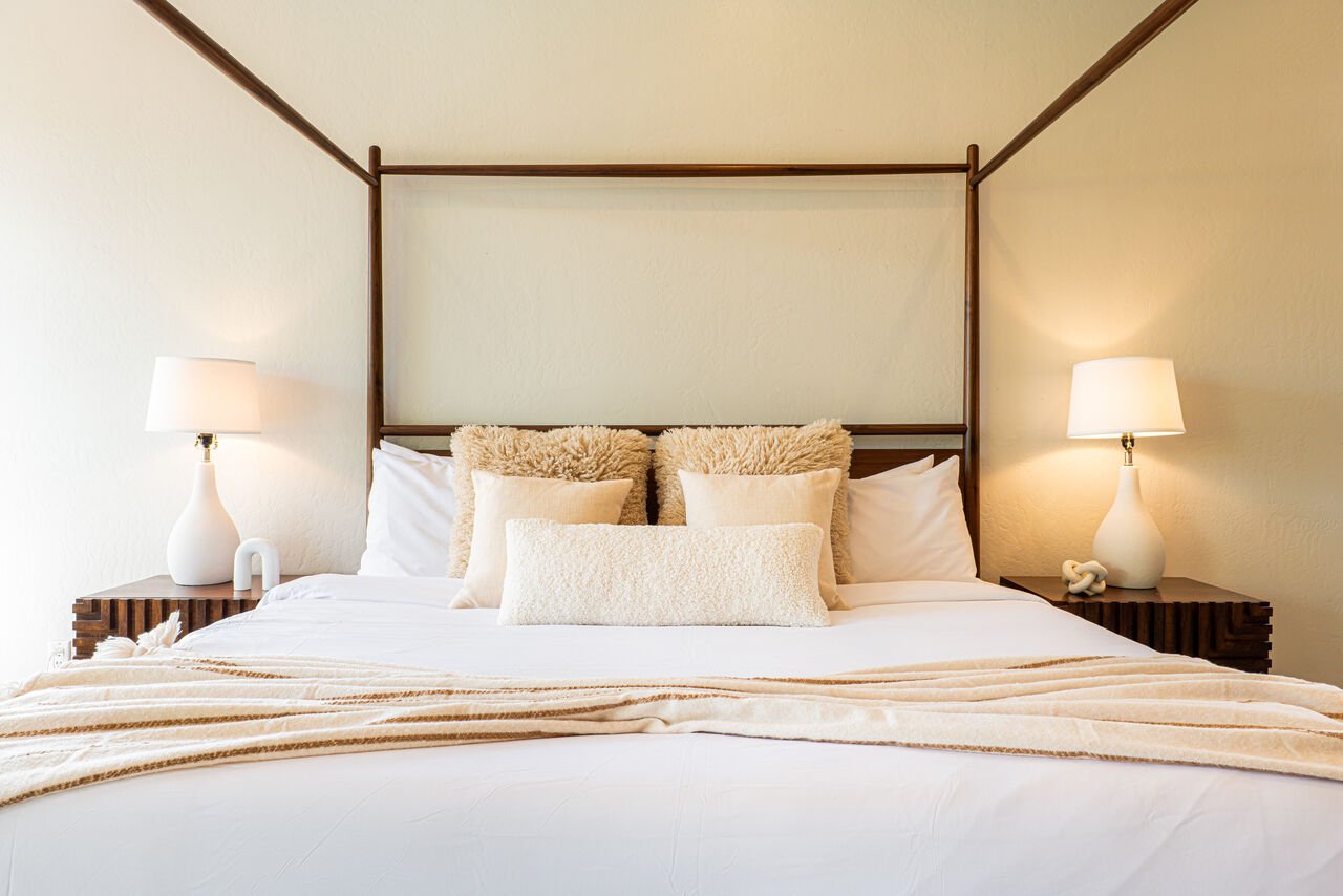 A neatly made bed with white sheets, beige and fluffy pillows, and a beige throw blanket, flanked by wooden nightstands with white table lamps in a softly lit room with cream-colored walls.