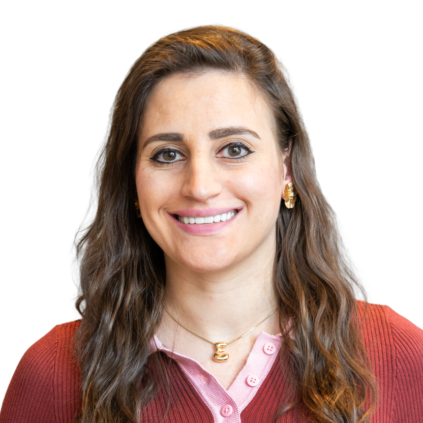 A woman with long wavy brown hair, light skin, and wearing a red sweater over a pink collared shirt, smiling at the camera.