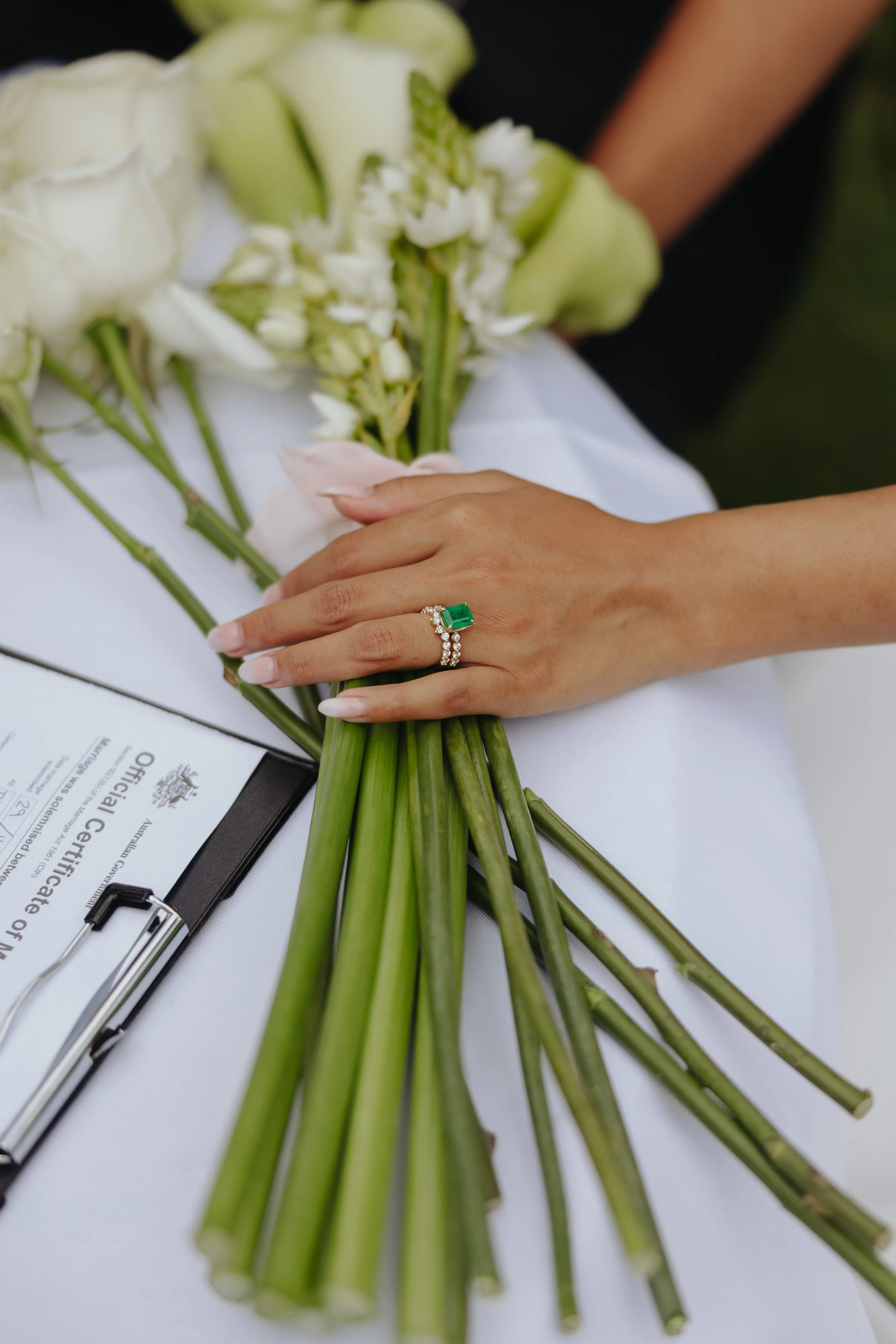 A bride holding a bouquet of peach roses and greenery, with a lace ribbon, standing beside a groom dressed in a dark suit, in front of a brick wall.