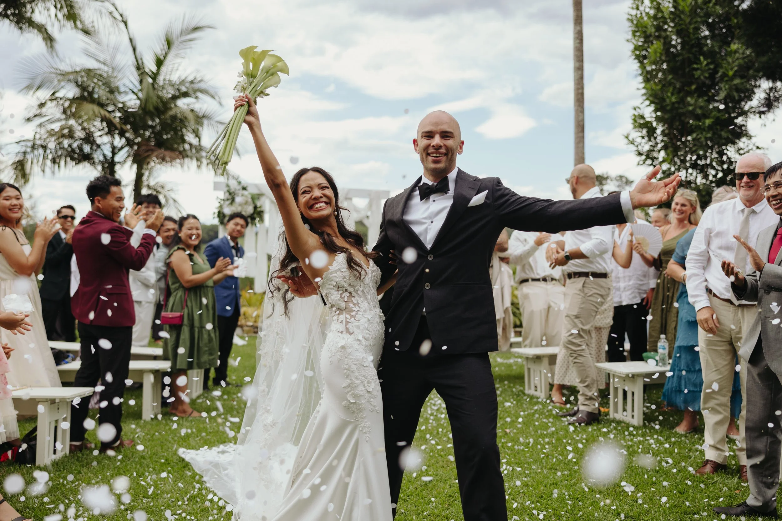 A bride raising her bouquet of flowers in the air in victory, standing beside a groom dressed in a dark suit, in the aisle after the wedding ceremony.
