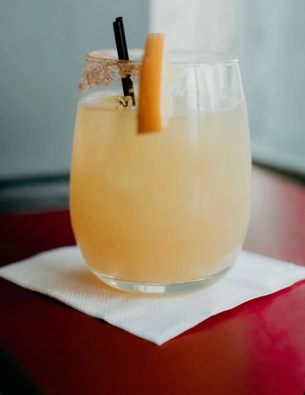 A glass of yellow cocktail with a slice of orange and a black straw on a white napkin.