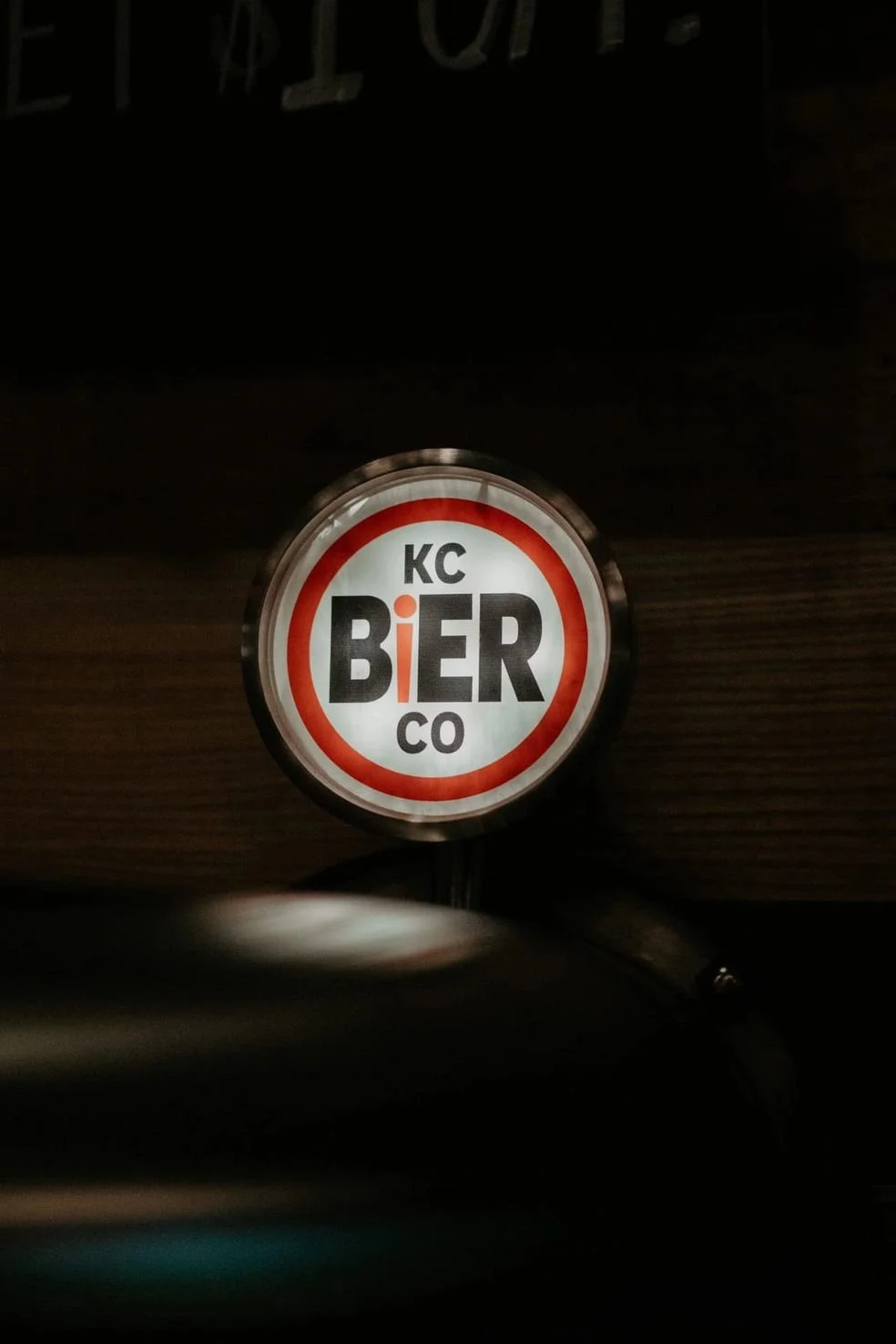 Illuminated circular sign with the text 'KC BIER CO' inside a red and black border, on a dark background.