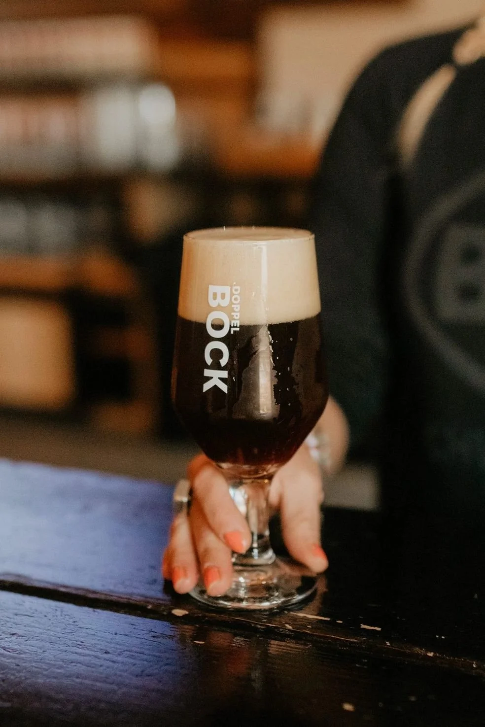 A hand holding a glass of dark beer with foam on top, on a wooden table.