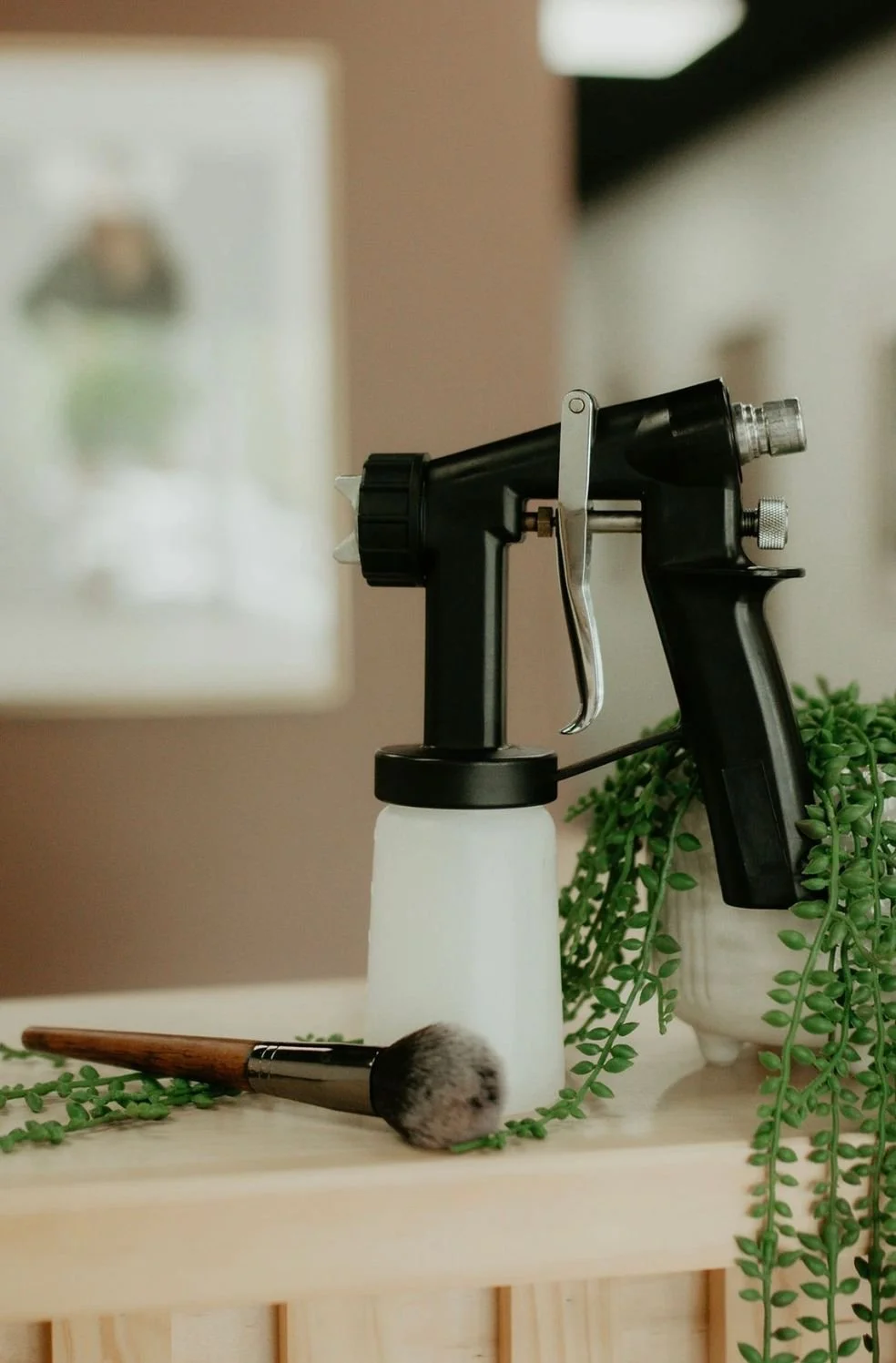 A black spray gun placed on a wooden surface, with a makeup brush and green plants nearby, and a blurred picture frame in the background.