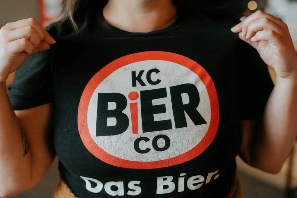 Person wearing a black T-shirt with a red, white, and black circular logo featuring the words "KC BIER CO" and "Das Bier."