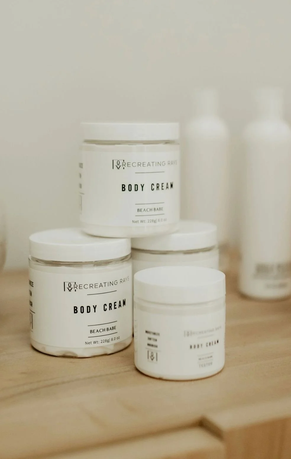 Several white jars of body cream labeled 'Recreating Rays' Beach Babe on a wooden surface with two white bottles in the background.