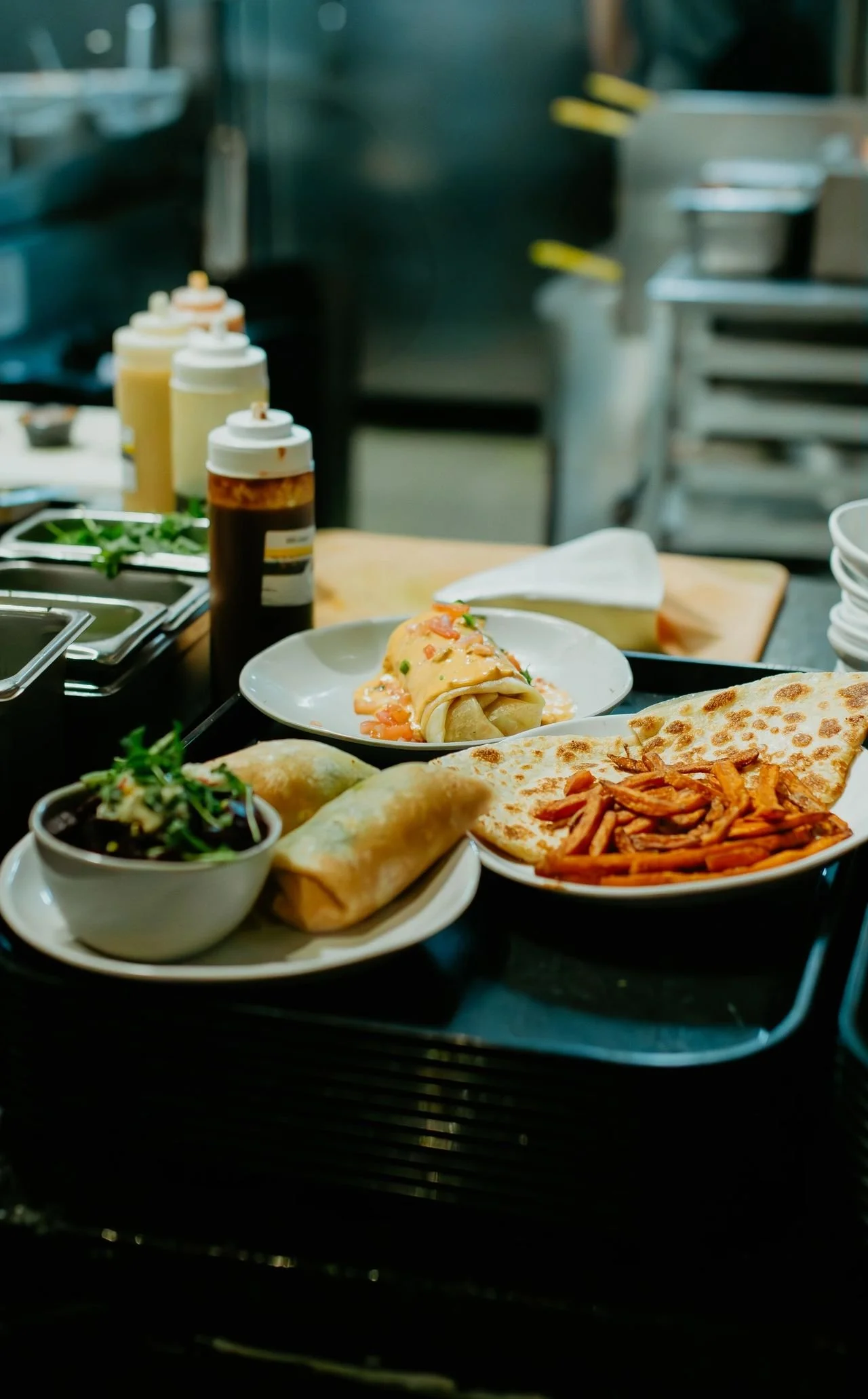A variety of Mexican dishes on a black counter, including a bowl of salad, two enchiladas, a quesadilla, and a plate of quesadillas with shredded chicken, with condiments in squeeze bottles in the background.