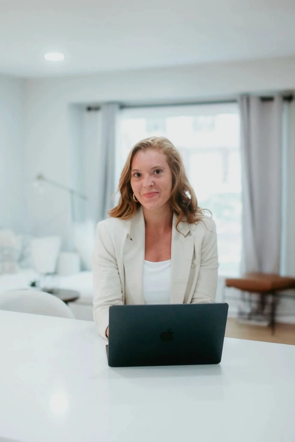 A woman with shoulder-length red hair, wearing a white blazer, sitting at a white table with a black laptop in front of her, in a bright and modern living room with large windows and light-colored curtains.