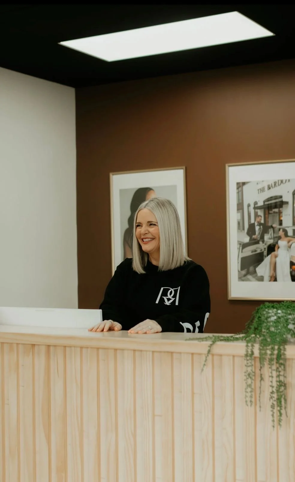 A woman with shoulder-length gray hair smiling while sitting at a wooden reception desk. She is wearing a black sweatshirt with a logo and is inside a room with framed photographs on the wall behind her.