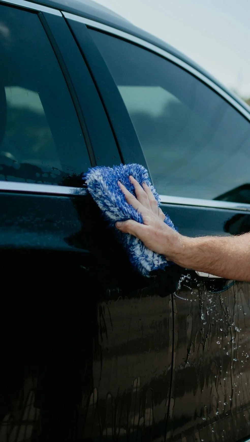 Person cleaning a black car with a blue and white microfiber cloth.