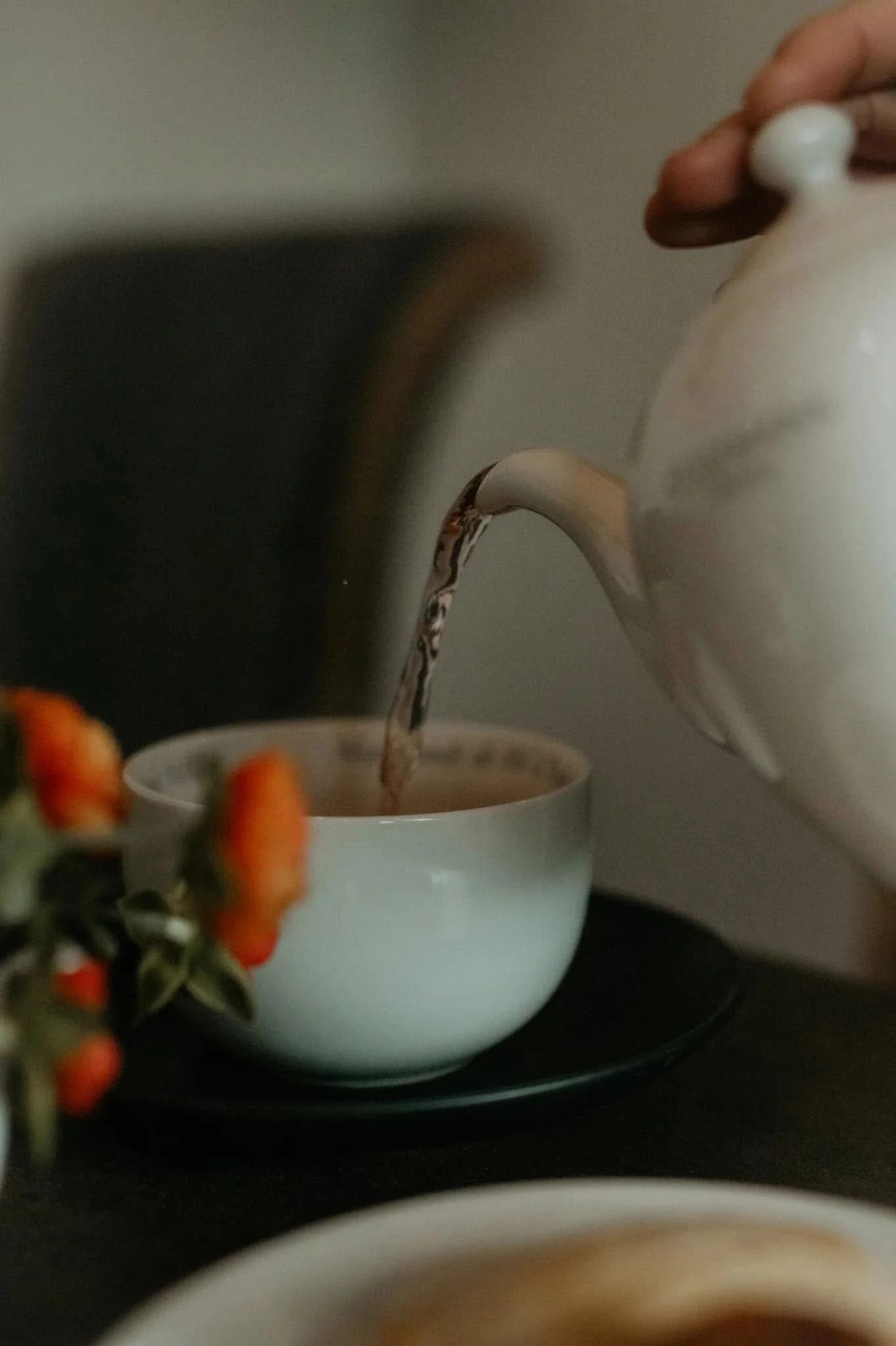 A white teapot pouring hot tea into a white teacup on a black tray, with a blurred bowl in the foreground and orange flowers on the left.