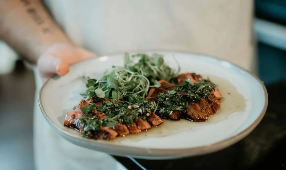 A person holds a white plate with sliced glazed salmon topped with herbs and a side of microgreens.