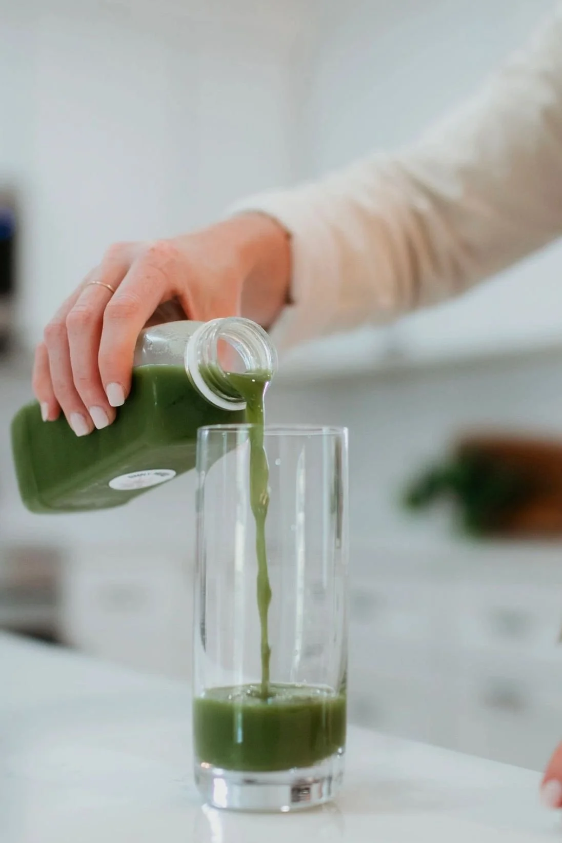 Person pouring green juice from a bottle into a tall glass.
