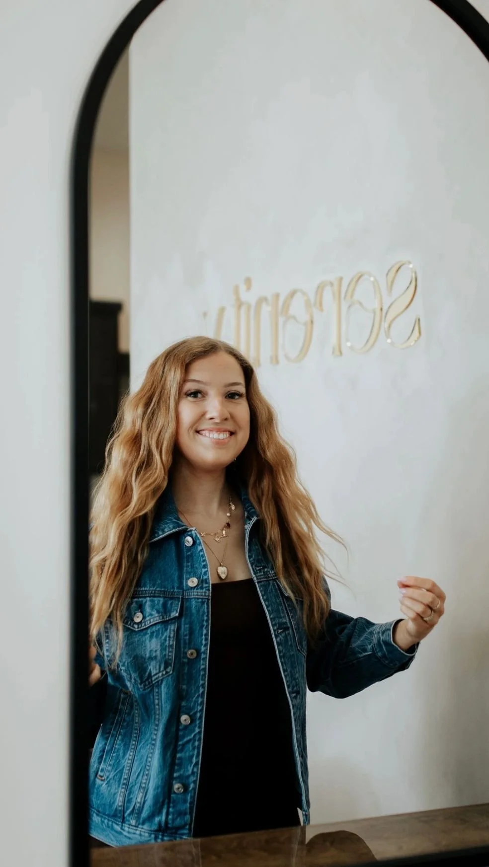 A smiling young woman with long, wavy, blonde hair taking a selfie in front of a mirror, wearing a denim jacket and layered necklaces.