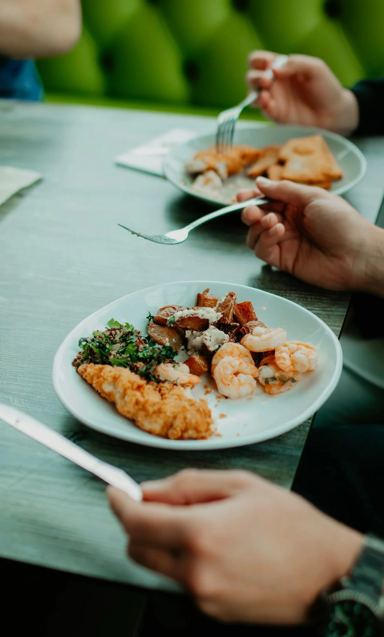 Close-up of a plate with shrimp, fried fish, and a mixed salad on a wooden table. In the background, a person is holding a fork and eating from a similar plate.