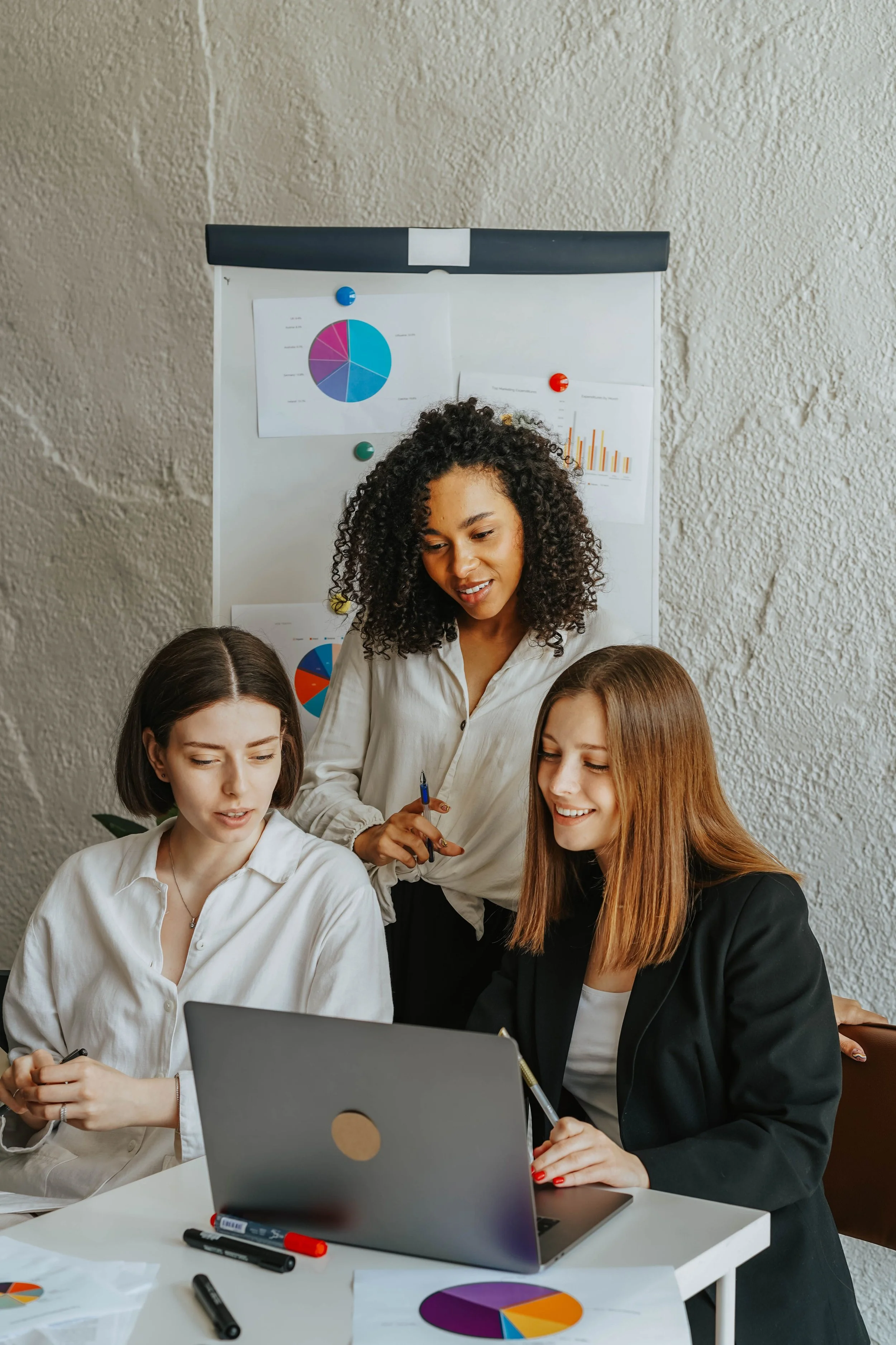 Group of women working together in a meeting room with charts on the wall, a laptop, and markers on the table.