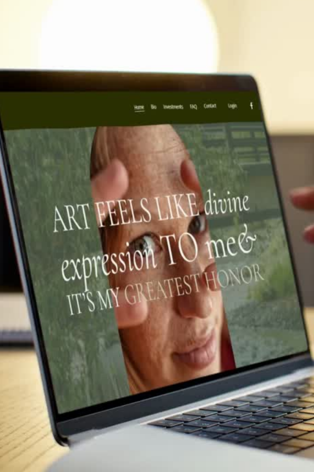 Close-up of a person holding a laptop showing a website with a woman's face and the text "ART FEELS LIKE divine expression TO me. IT'S MY GREATEST HONOR."