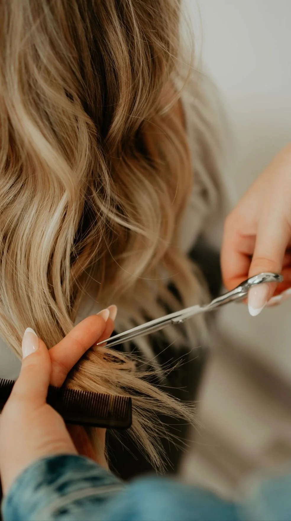 A person is cutting a woman's blonde wavy hair with scissors, holding a black comb.