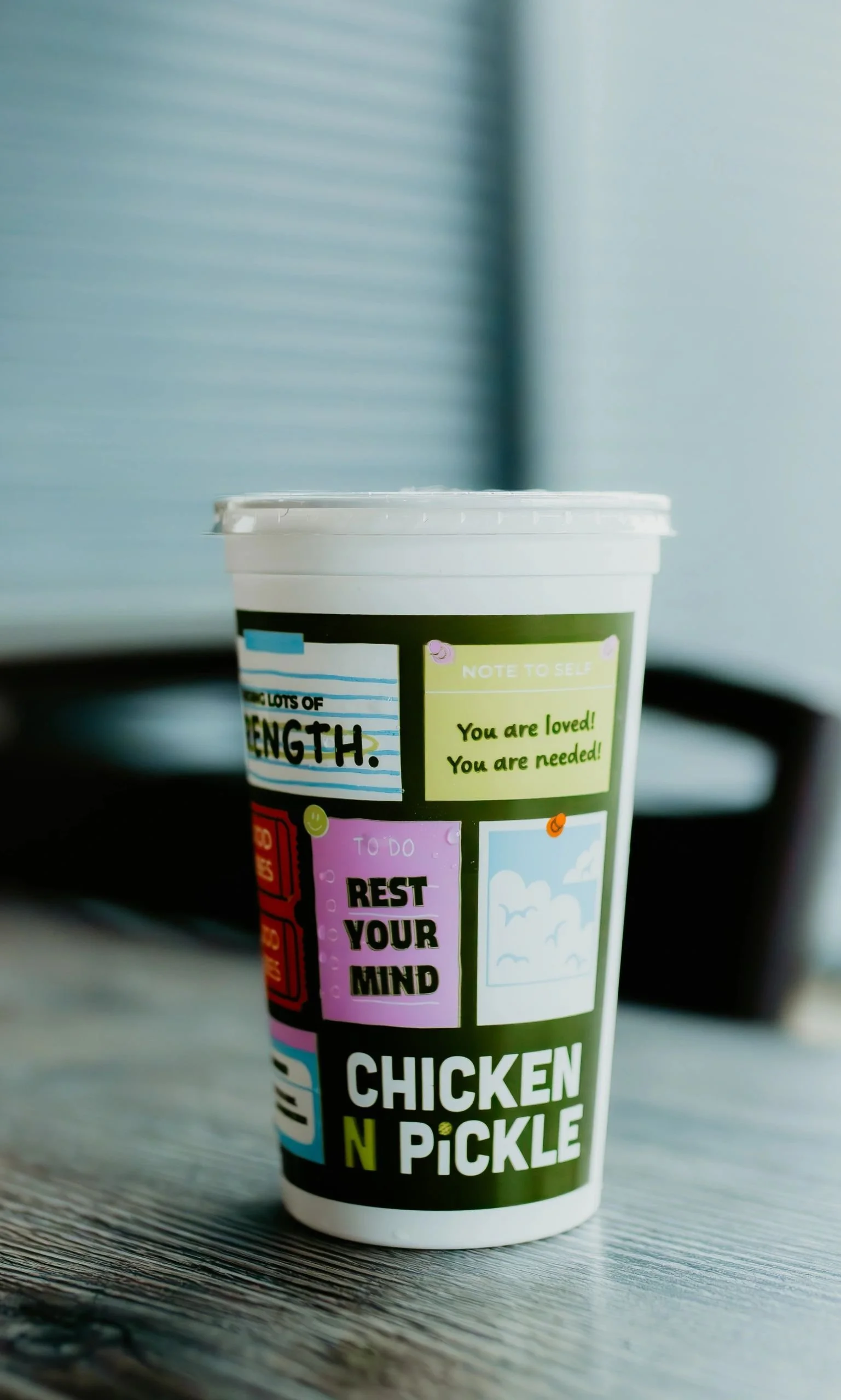 A paper cup with colorful motivational and positive messages, including 'Rest Your Mind', 'You are loved! You are needed!', and 'Lots of Strength', placed on a wooden surface.