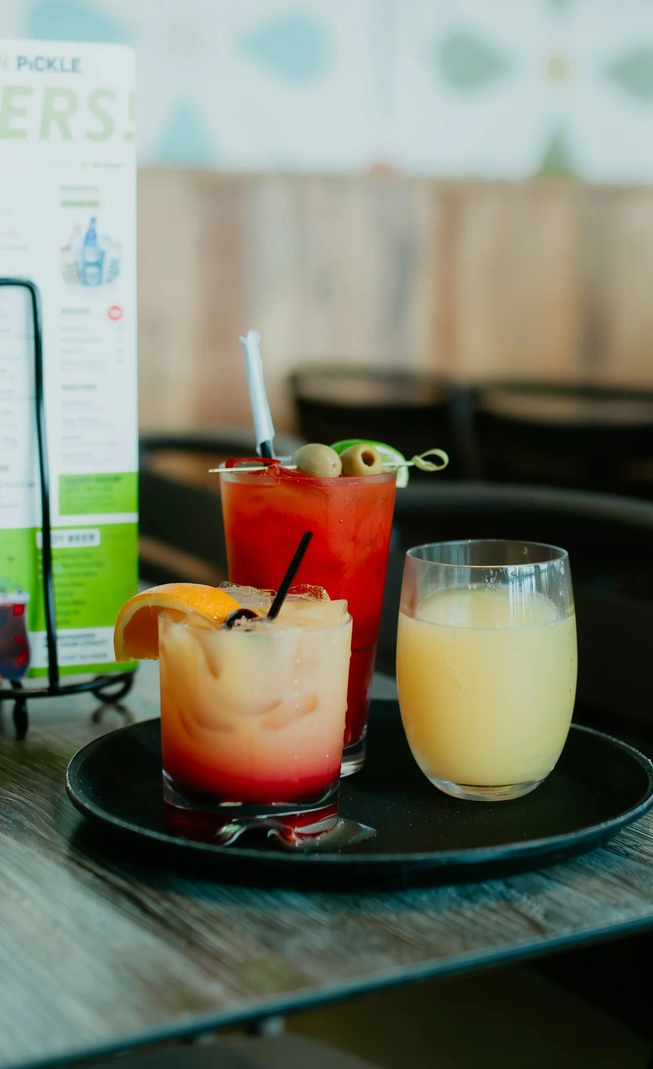 Three colorful cocktails on a black tray, including a red drink garnished with olives, a peach and red layered drink with an orange slice, and a yellow beverage, on a wooden table in a restaurant.