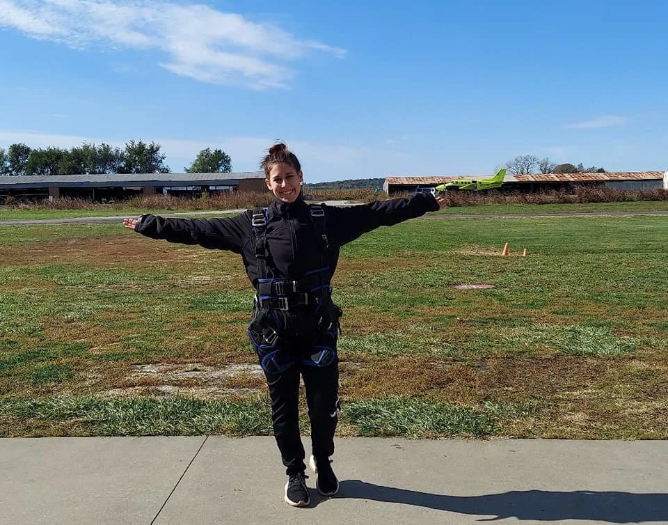 A woman in a skydiving jumpsuit standing on a sidewalk with arms outstretched, holding a small green airplane model in her right hand, at an outdoor airfield with a hangar and trees in the background on a sunny day.